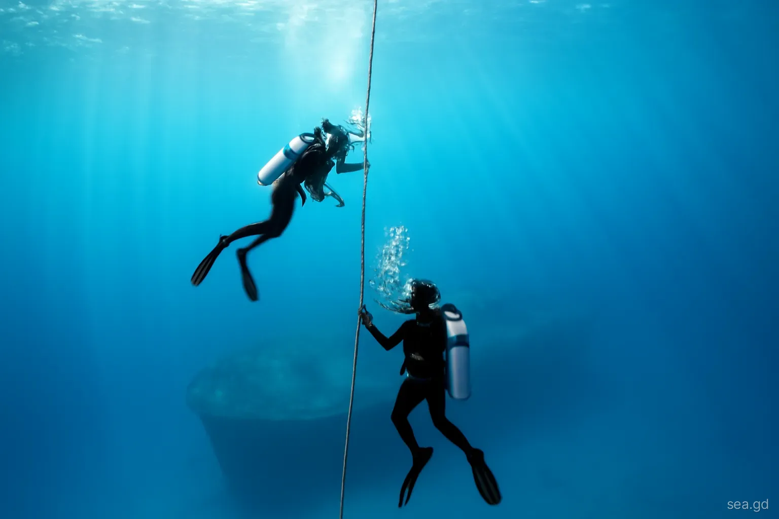 Two scuba divers descending a mooring line into deep blue water towards the shadowy outline of a large shipwreck off Grenada, view from above, sunbeams