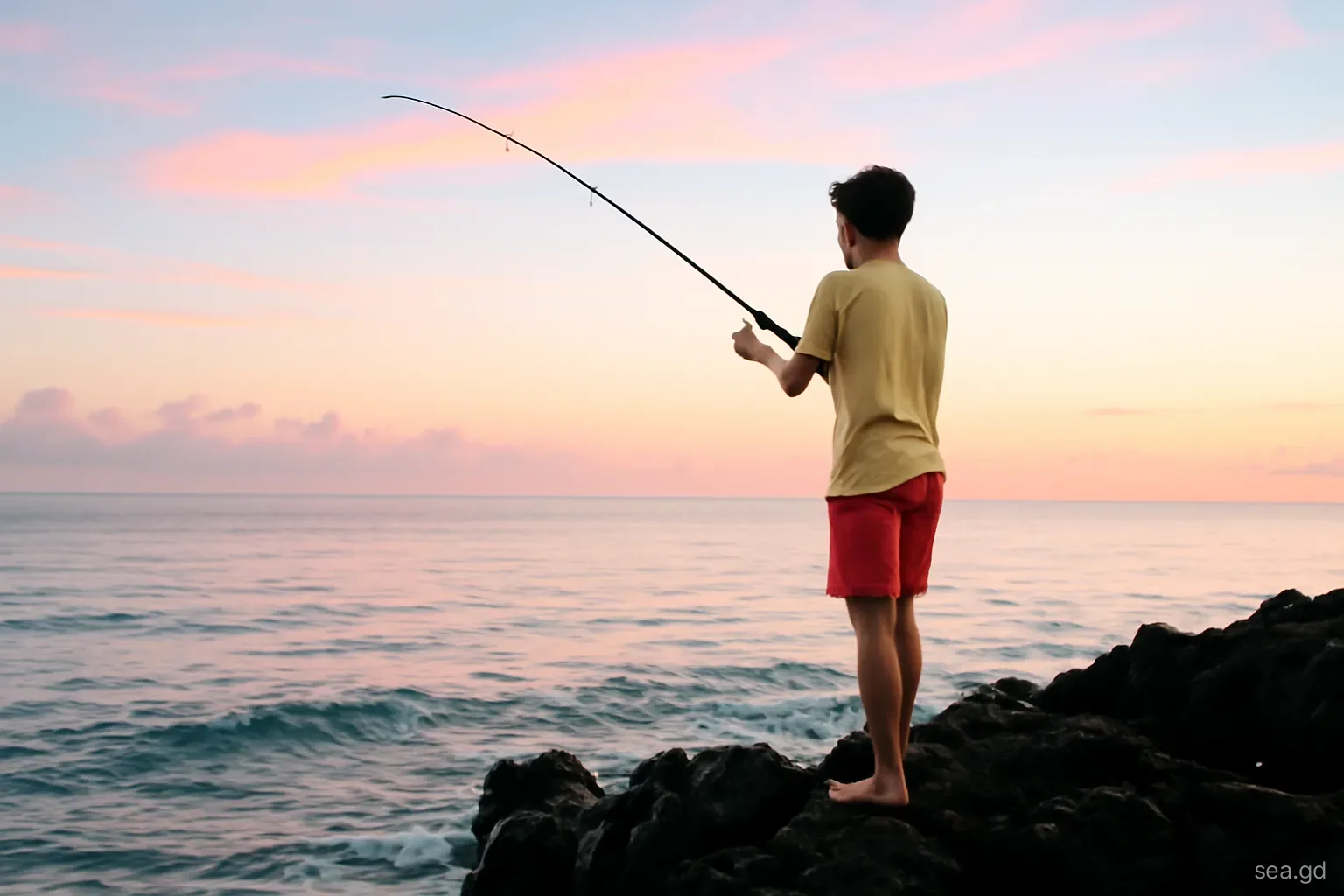 Teenage boy casting a spinning rod from dark volcanic rocks into a pink dawn sea on Grenada's south coast, silhouette from behind, gentle waves below