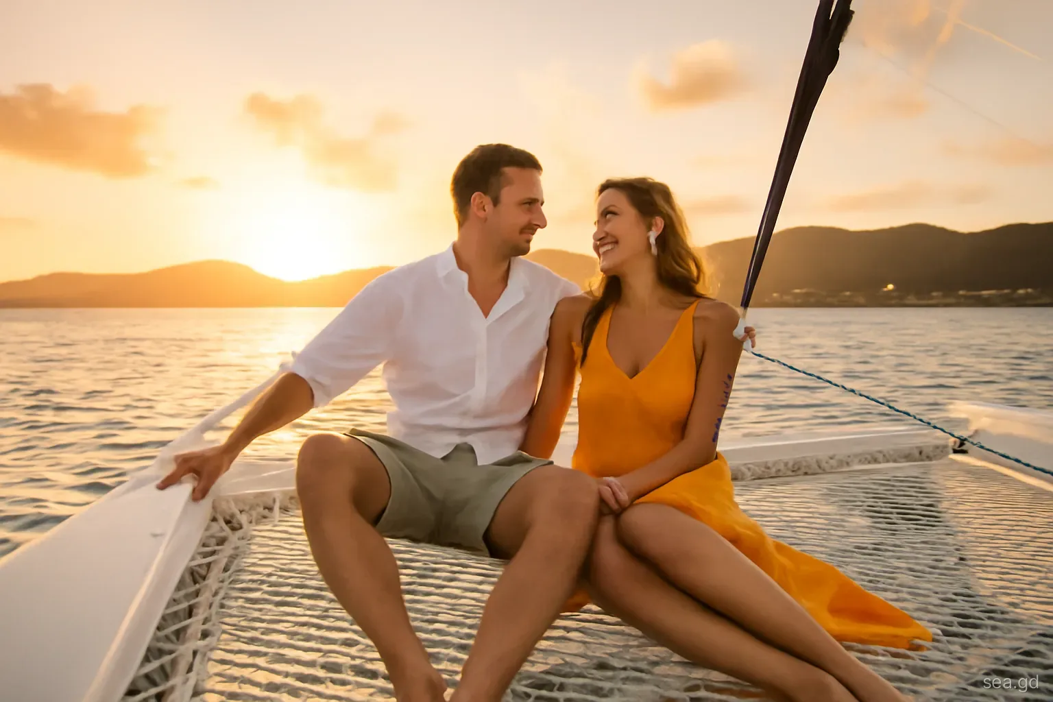 Couple seated on the bow net of a small catamaran at golden hour off Grand Anse, warm orange sky, silhouette of St George's hills, low angle from deck